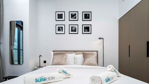 Quarto de hotel com cama, cabeceira, mesa de cabeceira, luminária, janela, espelho e parede com quadros.