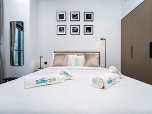 A hotel room with a bed, a headboard, a nightstand, a lamp, a window, a mirror, and a wall with framed pictures.