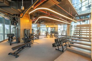 Un gimnasio moderno con diverso equipo de ejercicio, amplias ventanas y un techo alto con tuberías a la vista.