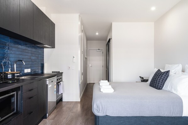 A modern hotel room with a kitchenette, a bed with white linens, and a view of the hallway. - City Centre Apartment on Hanover - Studio 1 (Dunedin)