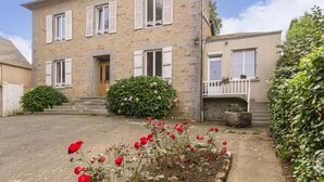 Une maison en pierre à deux étages, avec des volets blancs, une porte d’entrée et un petit porche doté d’une balustrade.