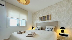 Ein ordentlich bezogenes Bett mit Handtüchern, ein Fenster mit Jalousien, eine Wand mit strukturierter Tapete und ein Nachttisch mit Lampe.