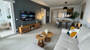 Ein modernes Wohnzimmer mit einem grauen Sofa, zwei runden Holztischen, Wandkunst im TV-Bereich und einem Essbereich mit Tisch und Stühlen.