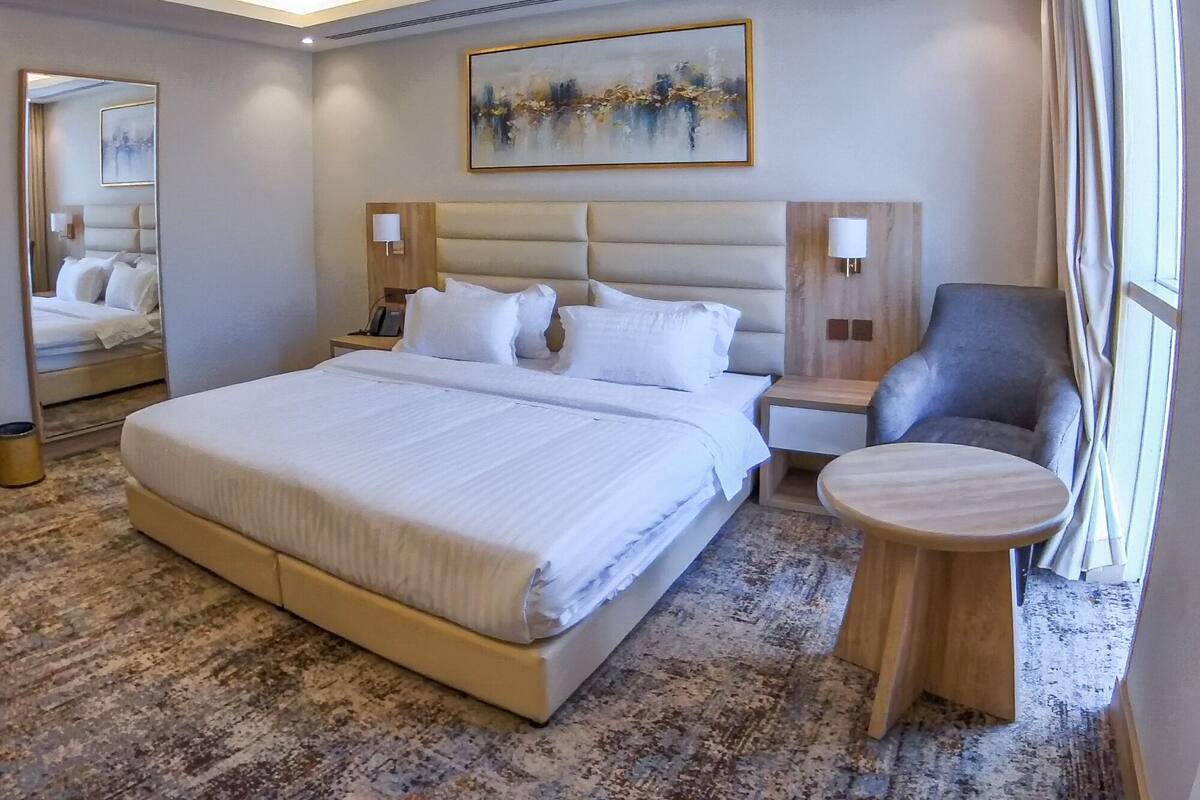 A modern hotel room with a large bed, a bedside table, a chair, a round table, and a painting on the wall.