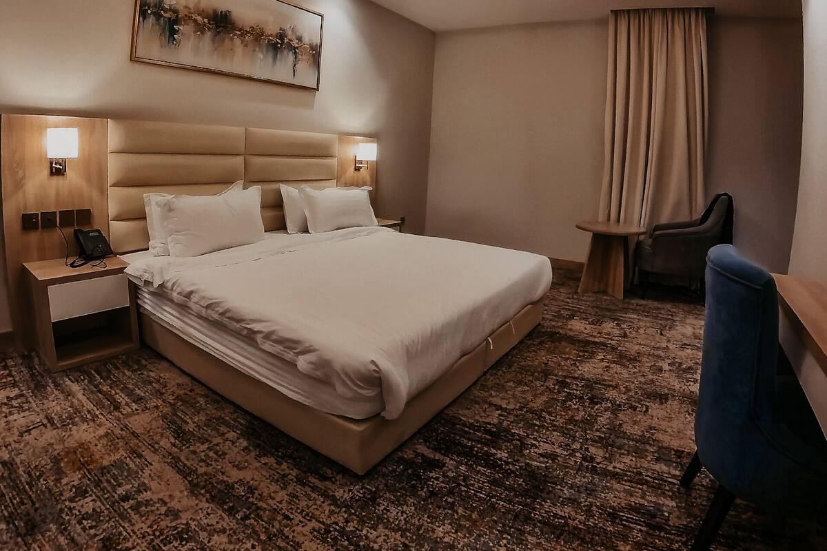 A hotel room with a large bed, two bedside tables with lamps, a desk with a chair, a painting on the wall, and a ceiling light.