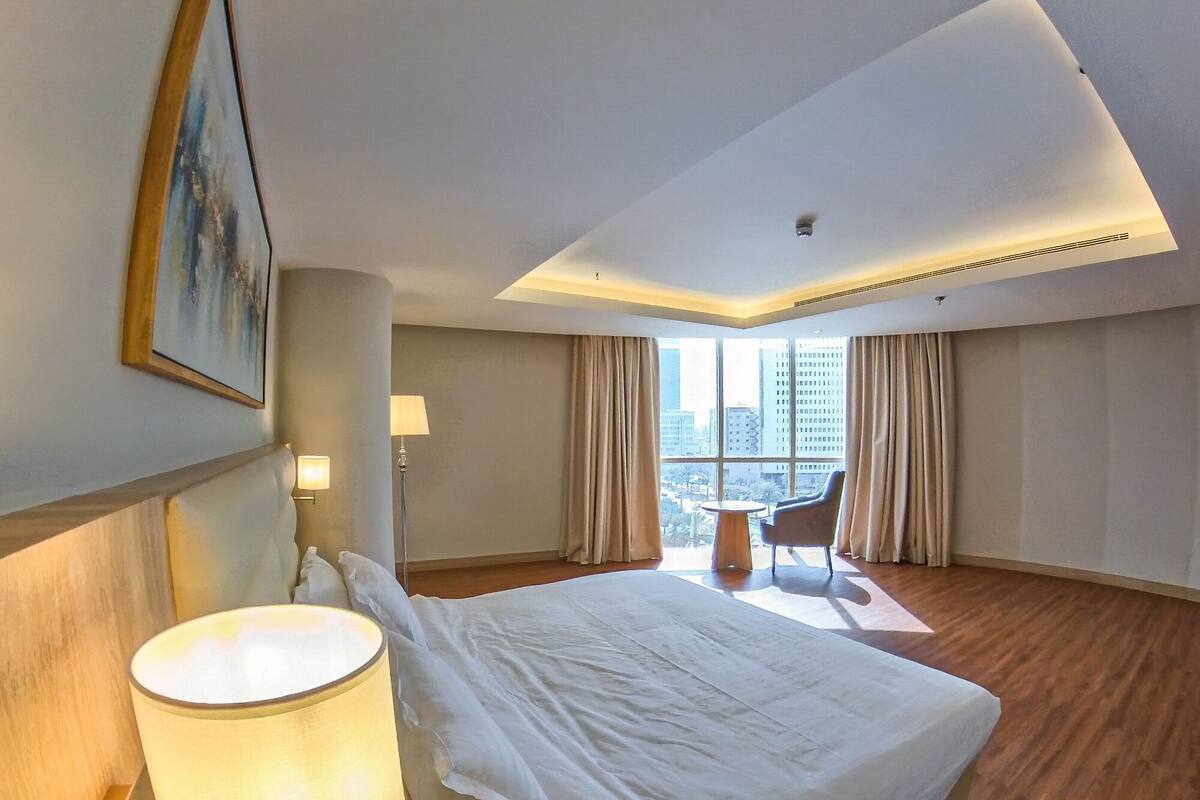 A hotel room with a large bed, a desk, a chair, and a view of the city through large windows.