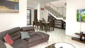 Una sala moderna con un sofá de cuero marrón, una zona de comedor con mesa y sillas, una escalera y un televisor montado en la pared.