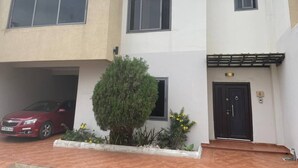 Un edificio de dos pisos con un coche rojo estacionado en la entrada, una puerta negra con una placa de números y un pequeño árbol en el jardín delantero.