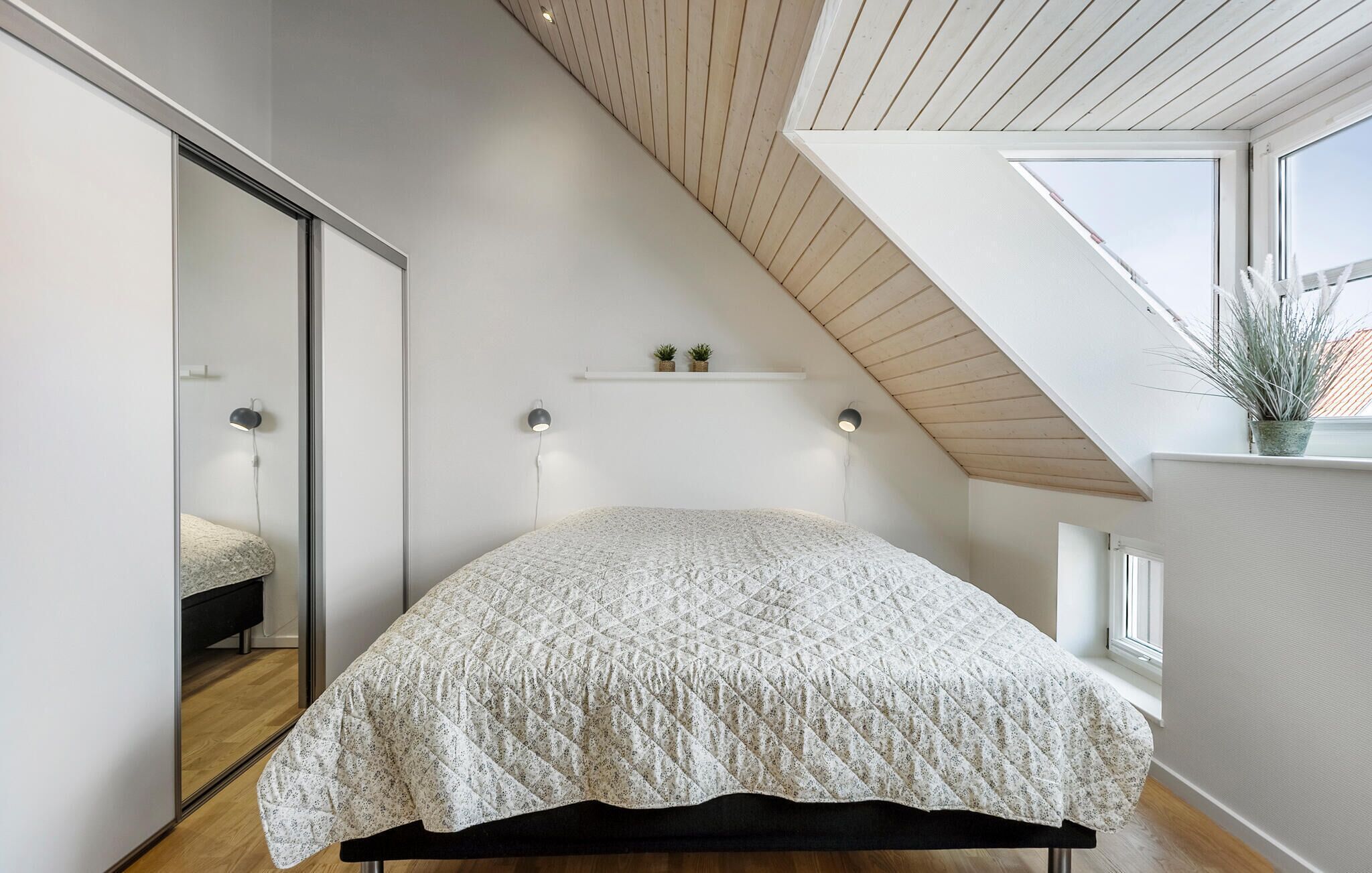 Un dormitorio con techo inclinado, una cama con colcha estampada, lámparas de noche, un pequeño estante con plantas y una ventana con una planta en el alféizar.