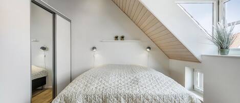 Un dormitorio con techo inclinado, una cama con colcha estampada, lámparas de noche, un pequeño estante con plantas y una ventana con una planta en el alféizar.