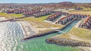 Zona residencial costera con casas alineadas a lo largo de la costa, un puente sobre un canal de agua y una vista distante de una colina con árboles.