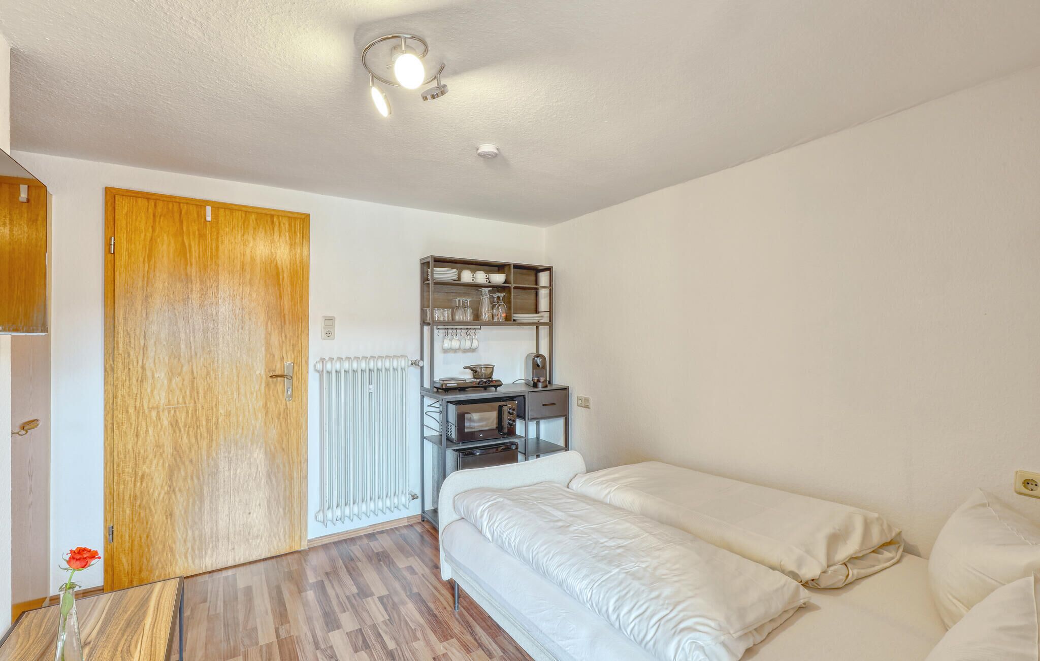 A bedroom with a wooden door, a radiator, a small kitchenette, and a bed.