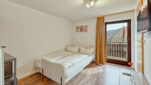 A small, single-bed room with a wooden floor, a window with a view of buildings, a small table with a vase, and a wall-mounted TV.