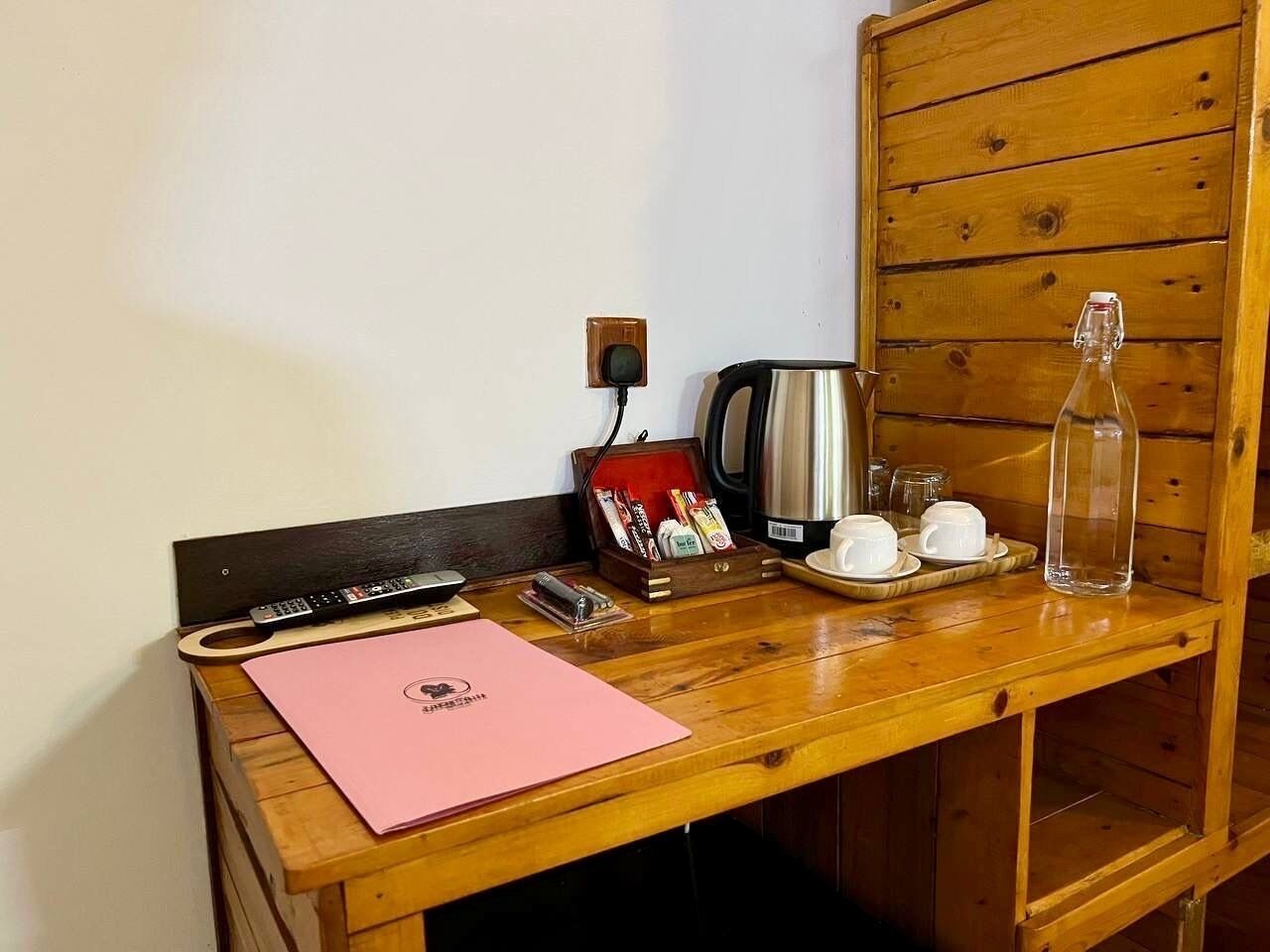 A wooden desk with a pink folder, a kettle, a bottle, and a box of tea bags.