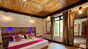 Un dormitorio con una cama grande, cabecera de madera y una pared decorativa de piedra. Se aprecia una ventana con cortinas y vistas al exterior con vegetación.