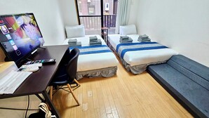 Une chambre d’hôtel avec deux lits, un bureau équipé d’un ordinateur et de télécommandes, une chaise et une fenêtre donnant sur des immeubles.
