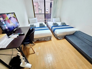 A hotel room with two beds, a desk with a computer and remote controls, a chair, and a window with a view of buildings. - Fuji Flat (Tokyo)