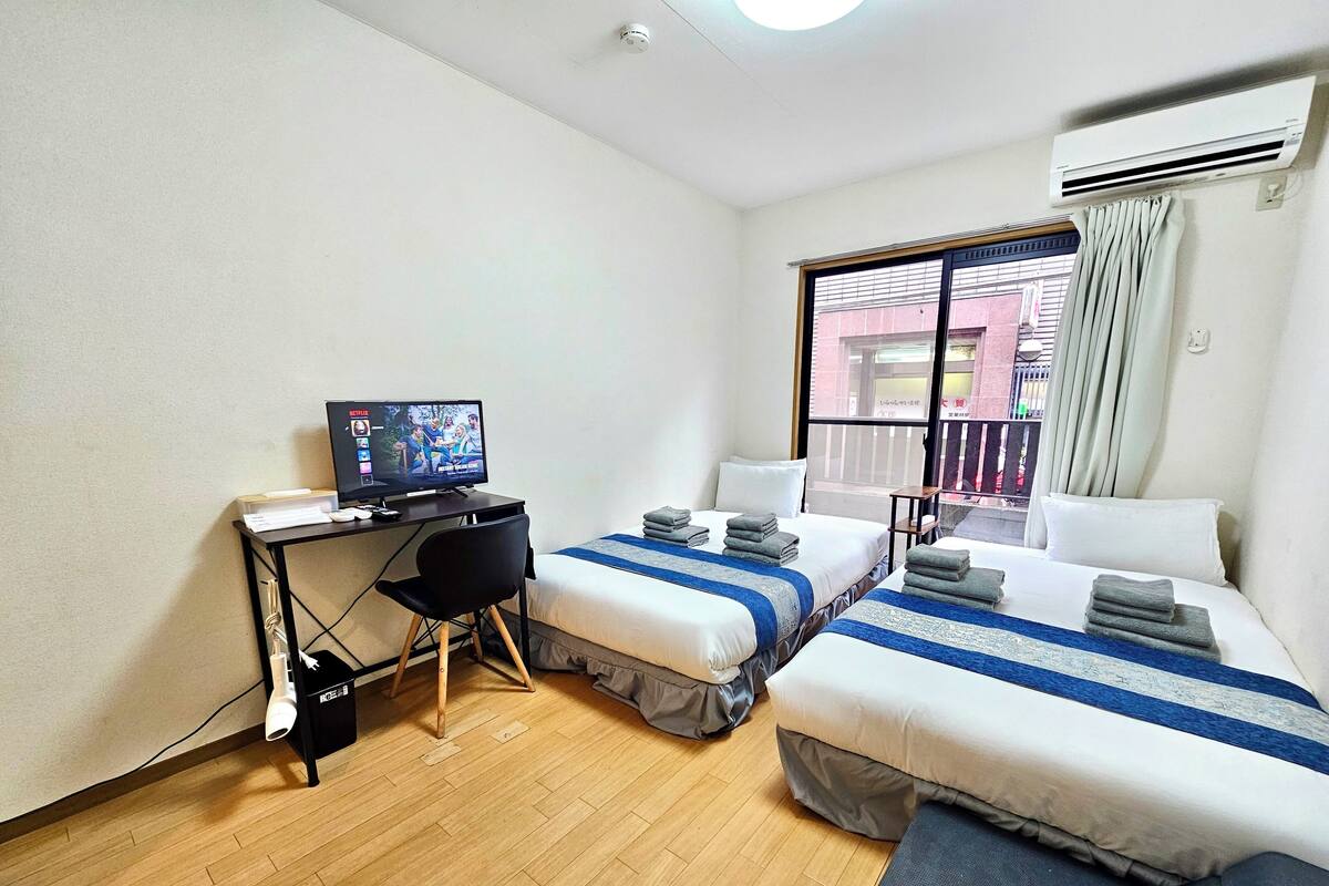 A hotel room with two beds, a desk with a computer, a chair, and a window with a view of buildings.