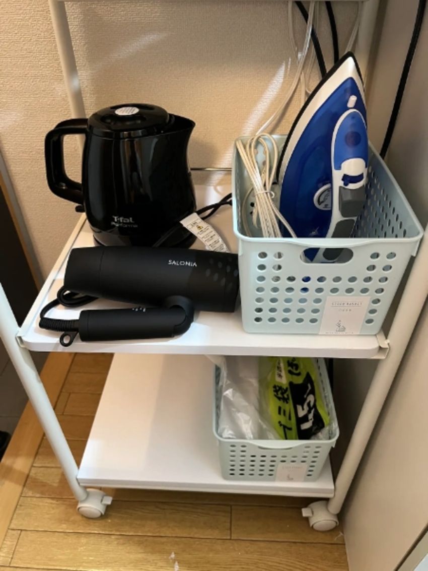 Una mensola in metallo bianco con un bollitore elettrico nero, un ferro da stiro blu e un altro ferro da stiro bianco sopra. In basso, due cesti bianchi, uno contenente una borsa verde e l'altro vuoto.
