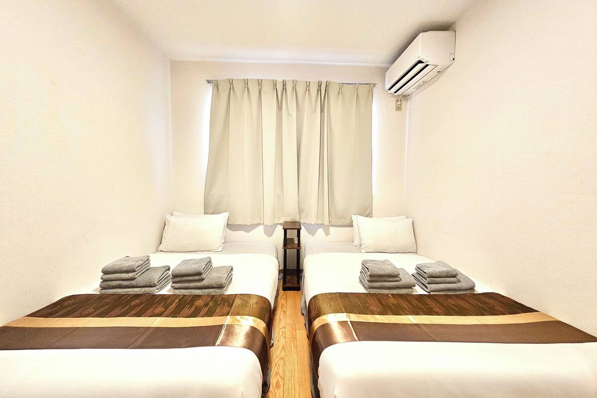 A hotel room with two beds, a window with curtains, an air conditioning unit, and a small table.
