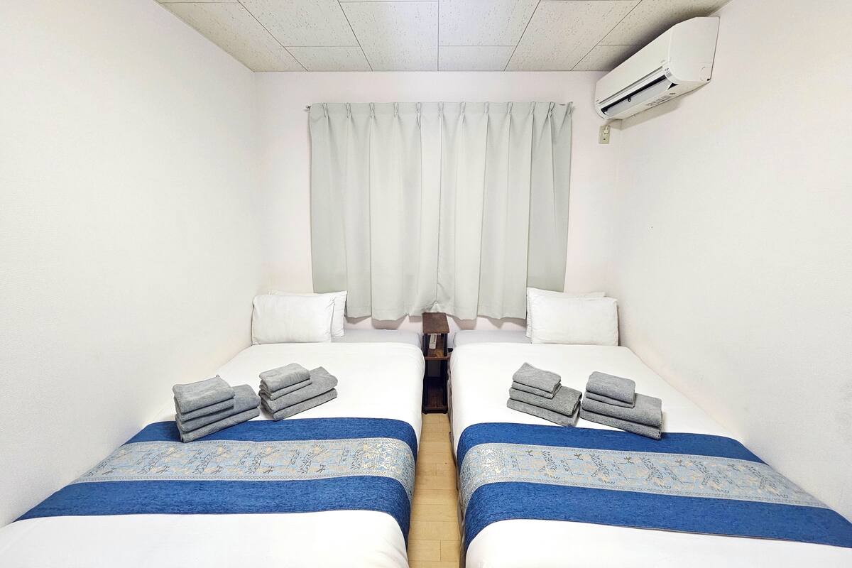 A small, clean room with two beds, each with a blue bedspread and pillows, and a set of towels on each bed.