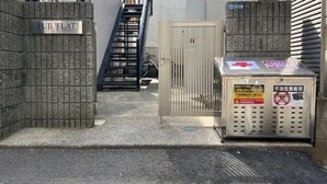 Une ruelle étroite, avec un portail portant l’inscription « FUJI FLAT », mène à un immeuble. On y trouve un escalier en métal et un vélo garé près du portail.