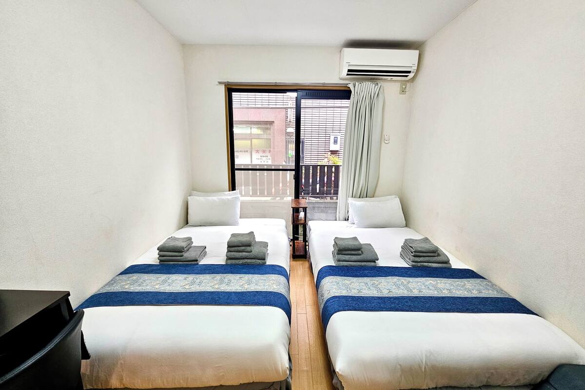 A room with two beds, a desk, an air conditioner, and a view through a window.