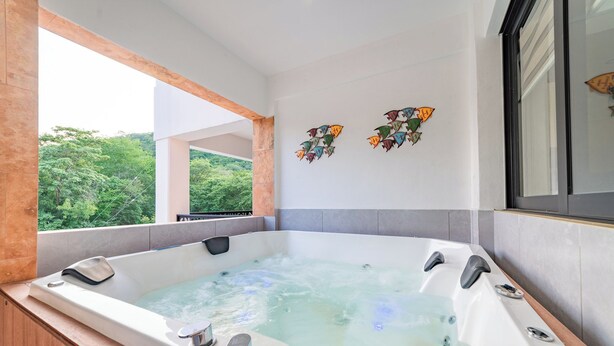 A modern bathroom with a large bathtub, a window with a view of greenery, and decorative wall art. - Centrally located condo with private plunge pool (Huatulco)