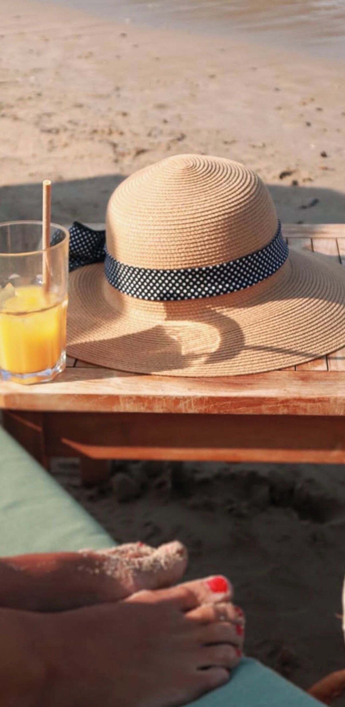 A straw hat with a polka dot band, a glass of orange juice, and a wooden table on a beach.