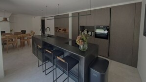 Uma cozinha moderna com uma ilha central, banquetas, uma área de refeições com cadeiras de madeira e eletrodomésticos embutidos.