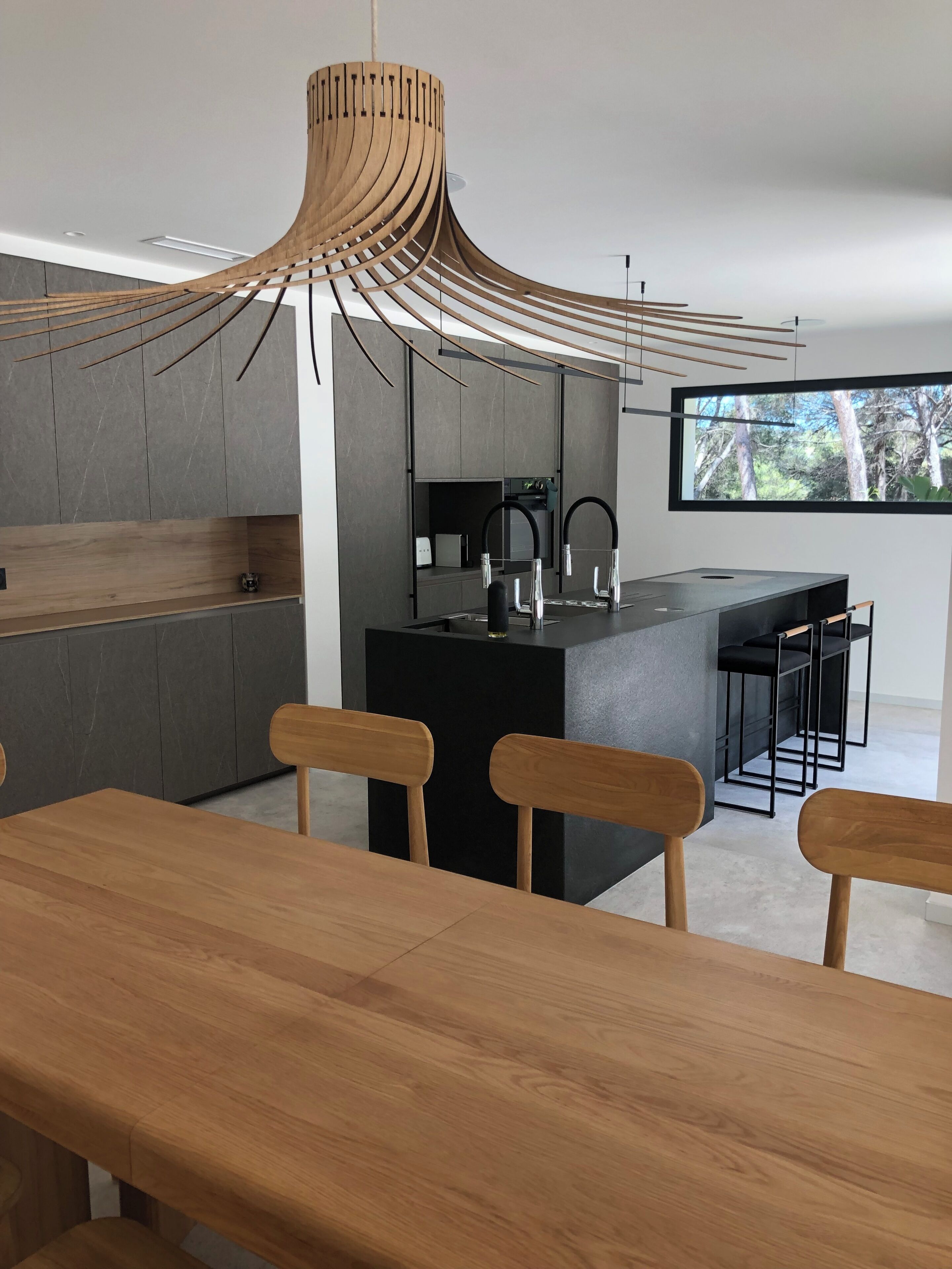Une cuisine moderne dotée d’un îlot central, d’un coin repas avec des chaises en bois et d’une grande fenêtre donnant sur la forêt.