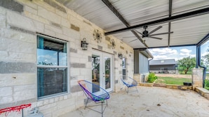A covered patio with stone flooring, two colorful wire chairs, a ceiling fan, and a view of a house in the background. - Countryside Texas Home w/ Fire Pit! (Richland Springs)
