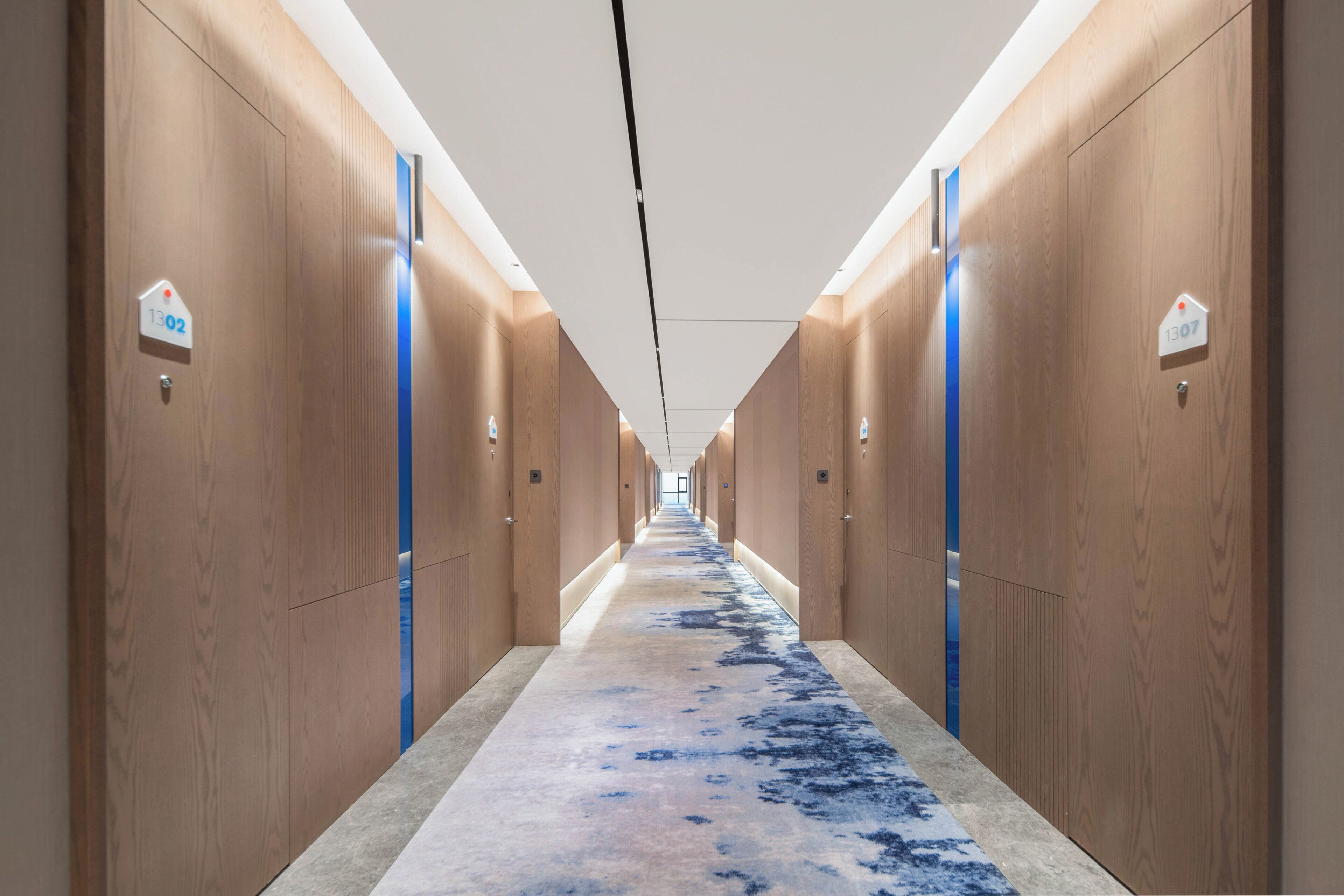 Um corredor de hotel com painéis de madeira, portas numeradas e um tapete com padrão azul e branco.