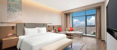 A modern hotel room with a large bed, a bench, a desk, and a large window offering a city view.