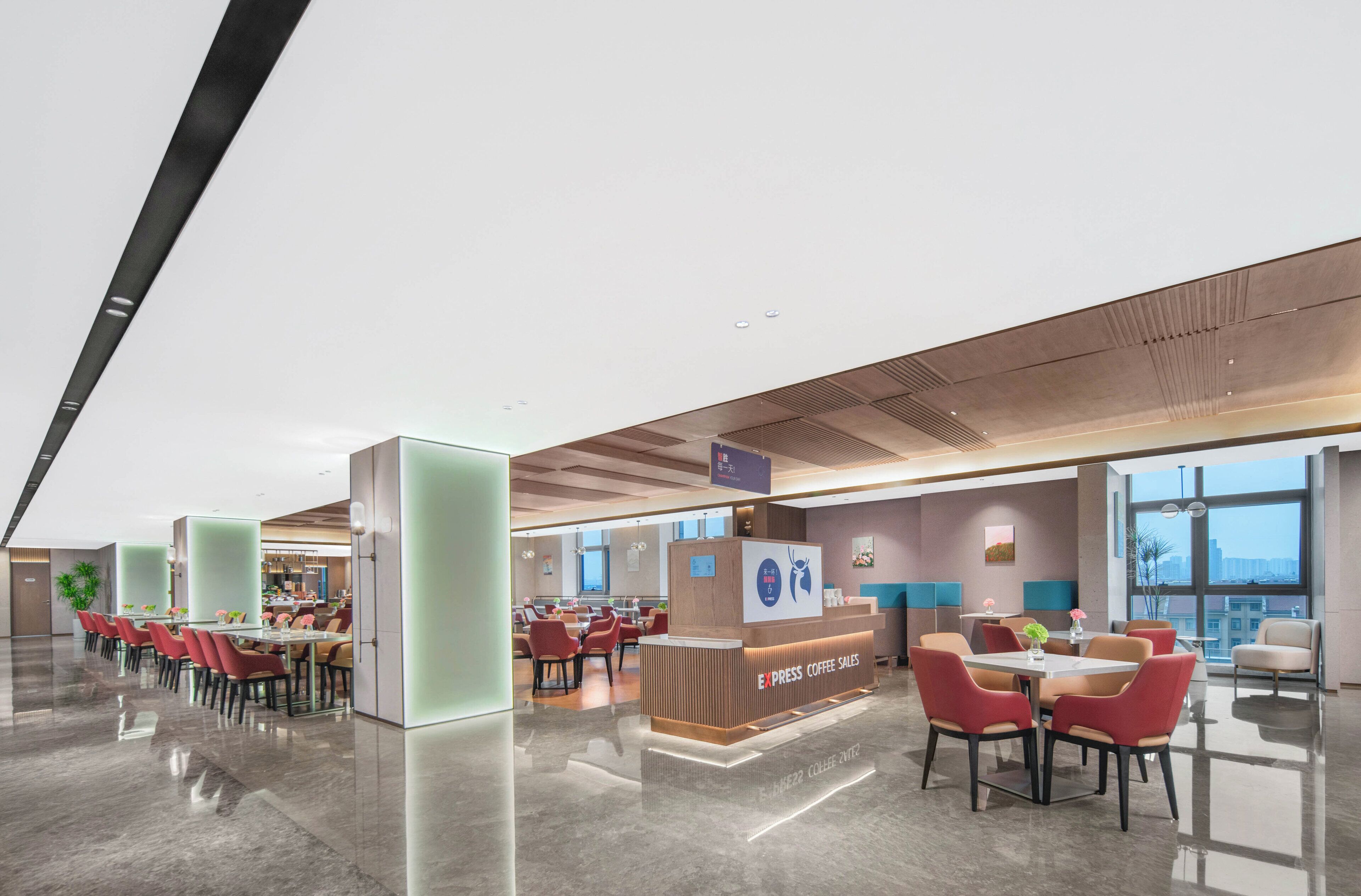 Un área amplia de cafetería con una recepción, diversas opciones de asientos y grandes ventanales con vistas.
