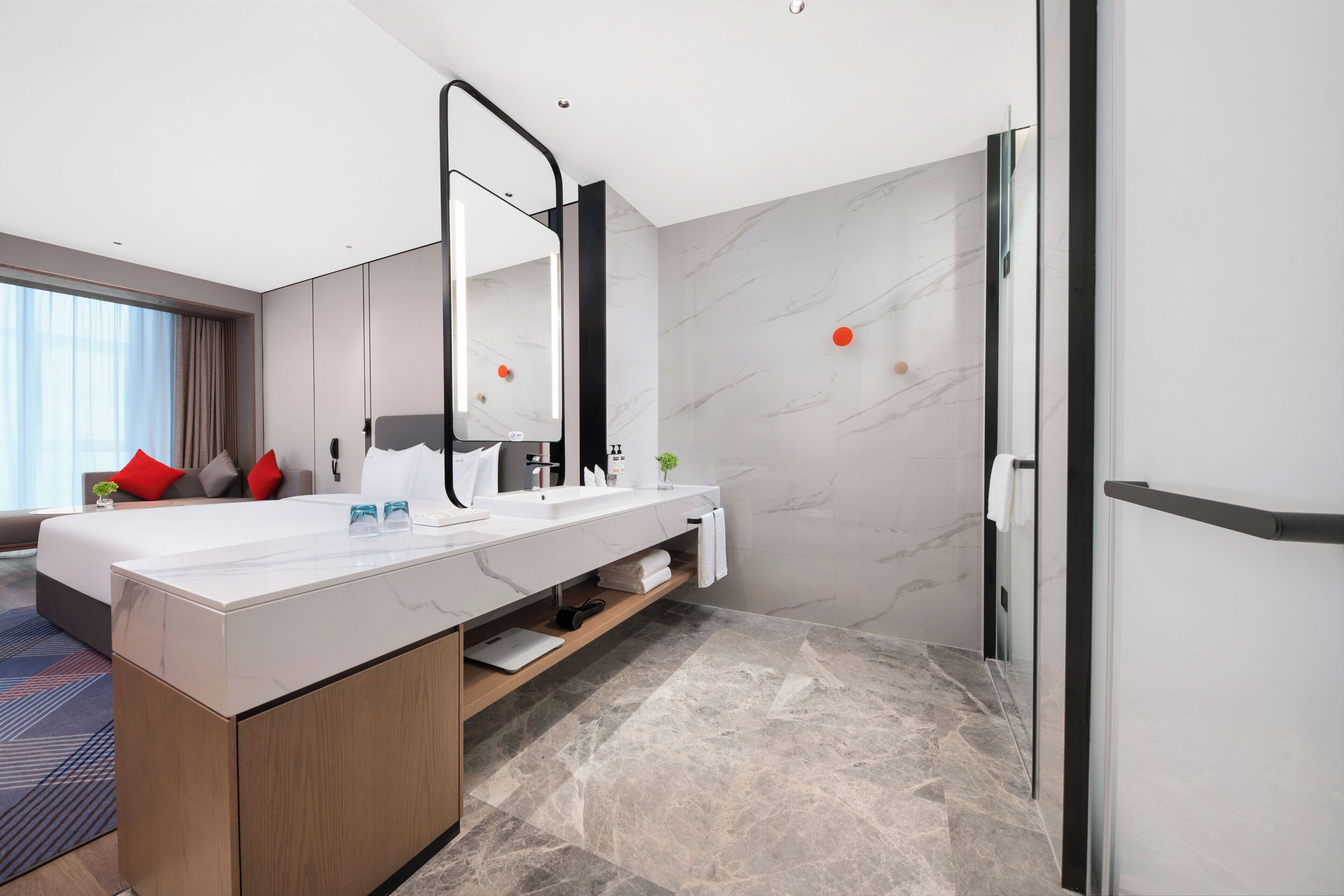 A modern hotel room with a large bed, a double sink vanity, a large mirror, and a shower area.