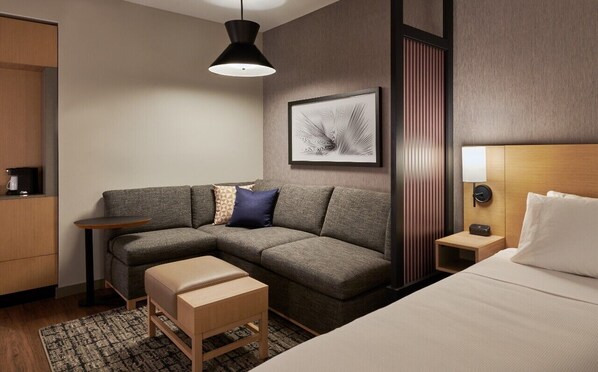 A modern hotel room with a sectional sofa, a small table, a bedside table with a lamp, a bed, and a framed artwork on the wall.