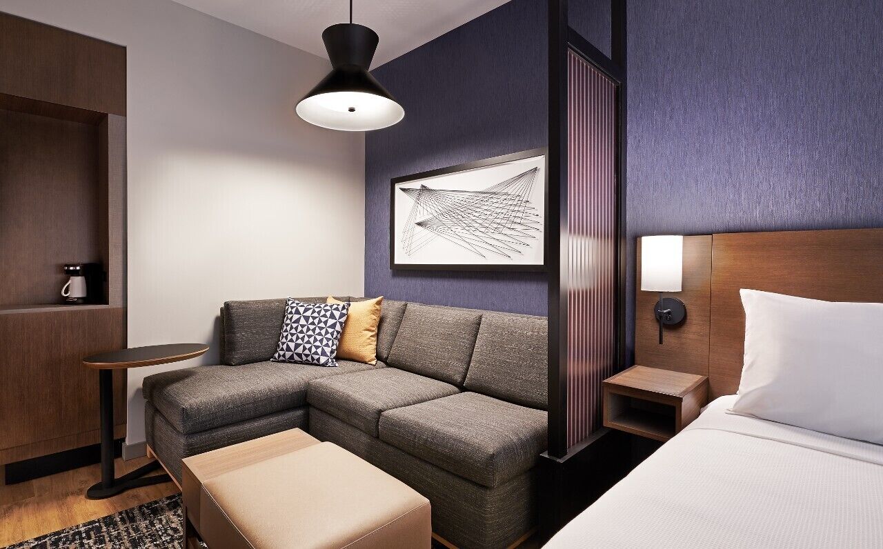 A modern hotel room with a sectional sofa, a bed, a small table, and a wall-mounted artwork.