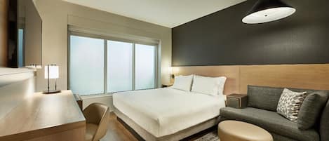 A modern hotel room with a large bed, a desk with a lamp, a sofa, and a large window providing natural light.