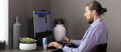 A person seated at a desk with a computer, a can of drink, and a potted plant.