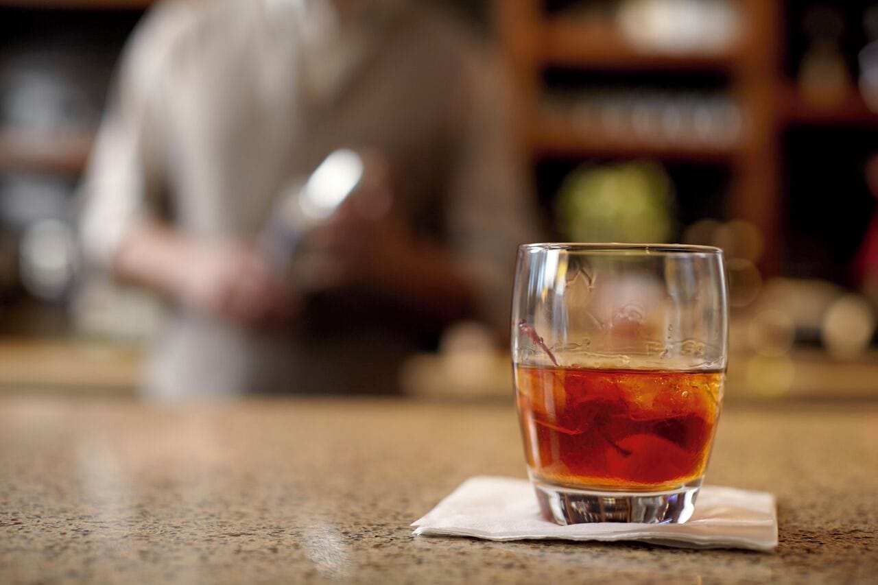 A glass of amber-colored beverage on a table with a blurred background of a person and shelves.