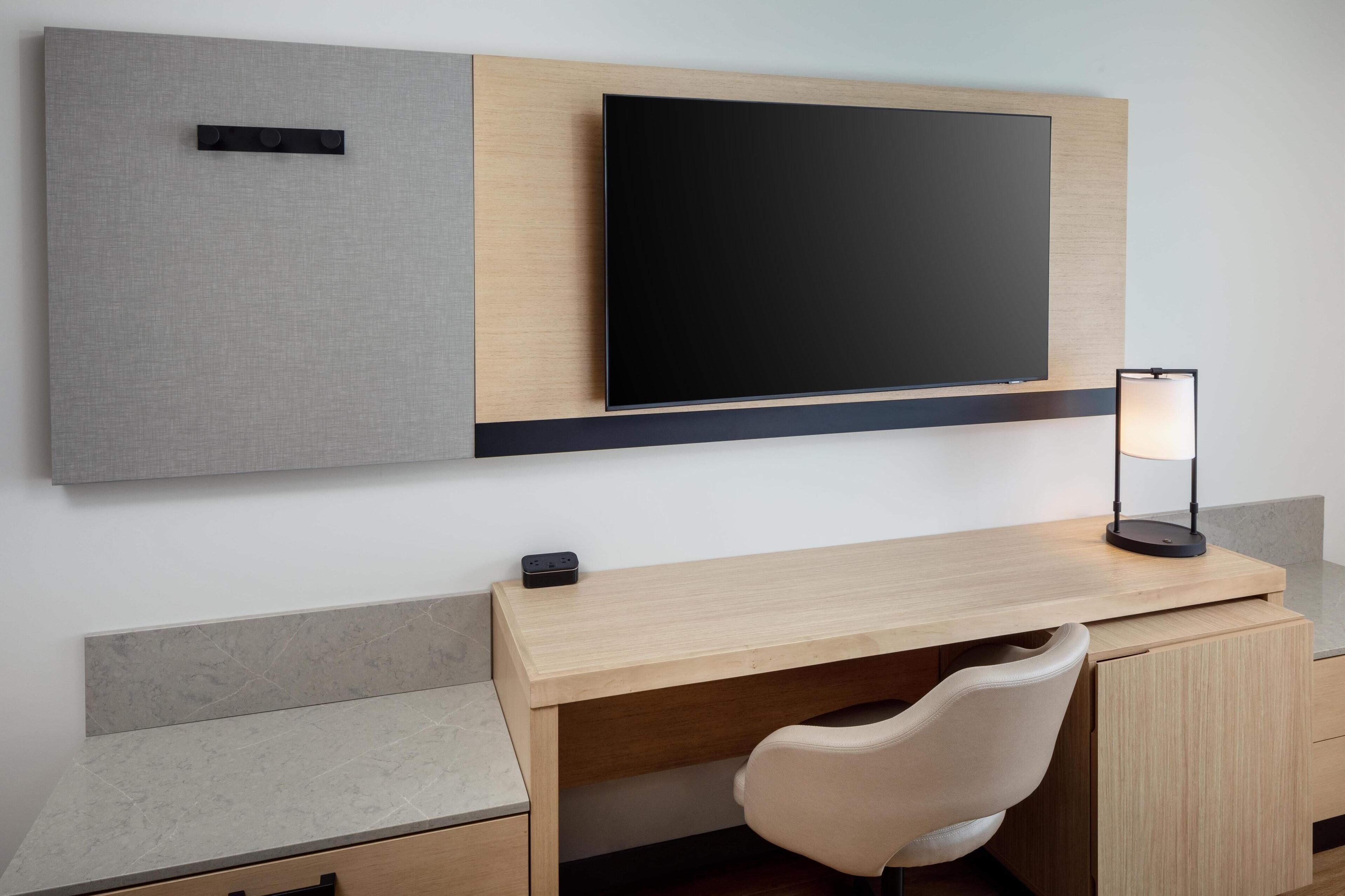 Ein modernes Hotelzimmer mit einem Holztisch, einem flachbildfernseher an der Wand, einer Schreibtischlampe und einem gepolsterten Stuhl.