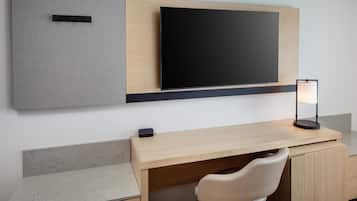 Una habitación de hotel moderna con un escritorio de madera, un televisor de pantalla plana montado en la pared, una lámpara de escritorio y una silla acolchada.