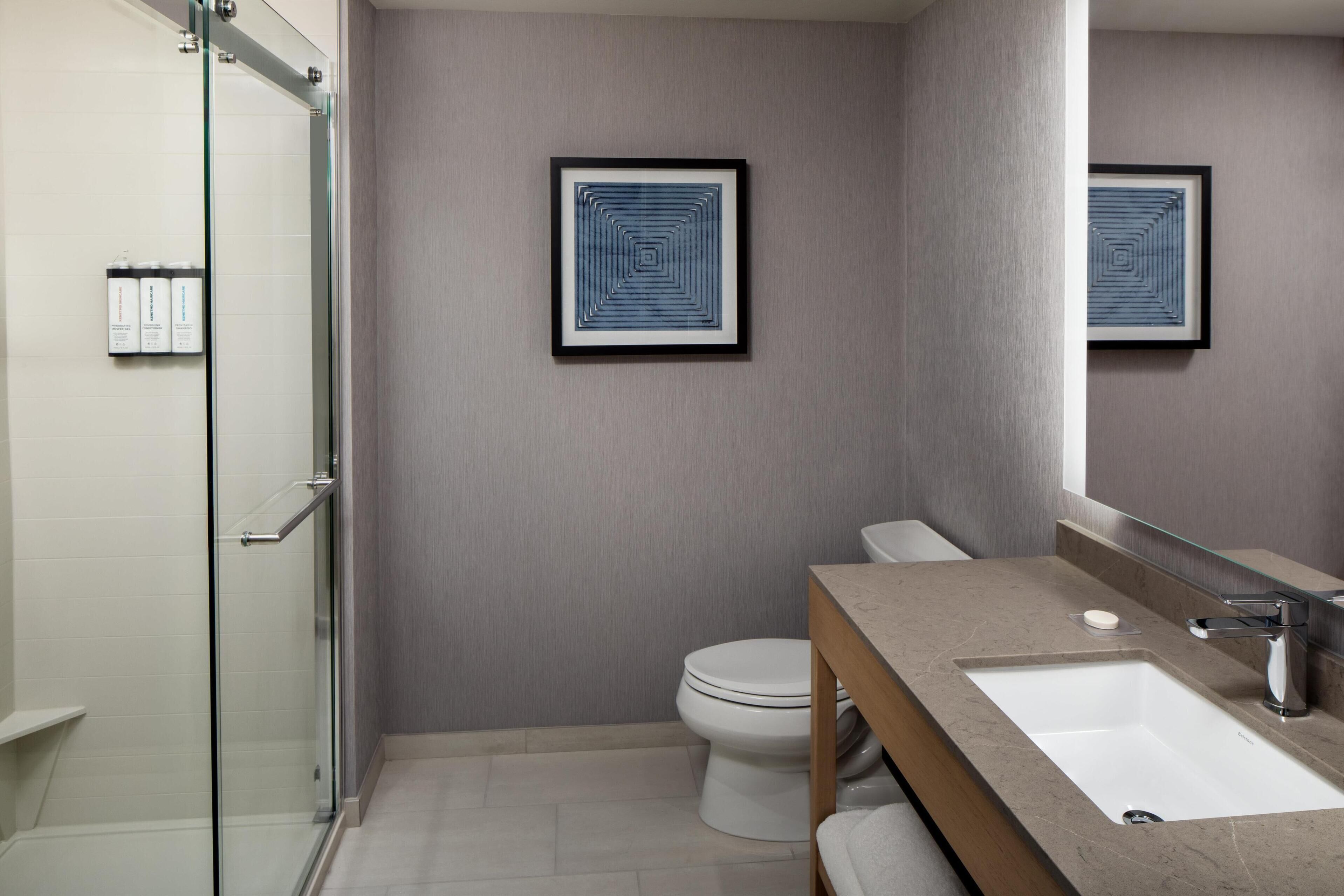 A modern bathroom with a glass shower enclosure, a white toilet, a beige vanity with a sink, and two framed wall art pieces.