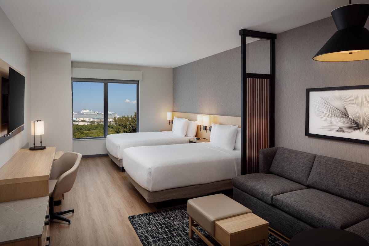 Ein modernes Hotelzimmer mit zwei Betten, einem Schreibtisch, einer Sitzecke und einem großen Fenster mit Blick auf die Stadt.