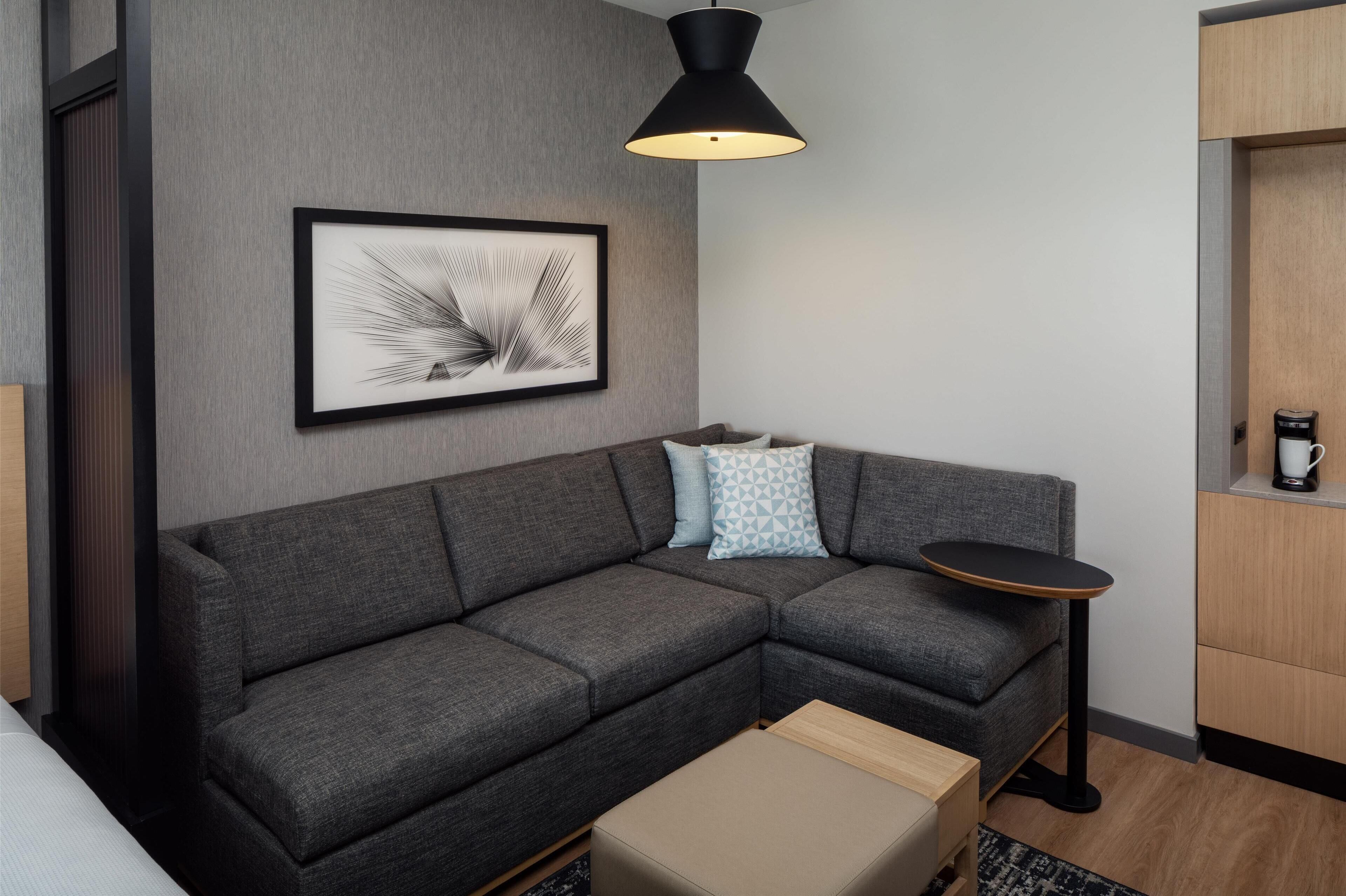 A modern living room with a gray sectional sofa, a small round table, a coffee machine, and a framed artwork on the wall.