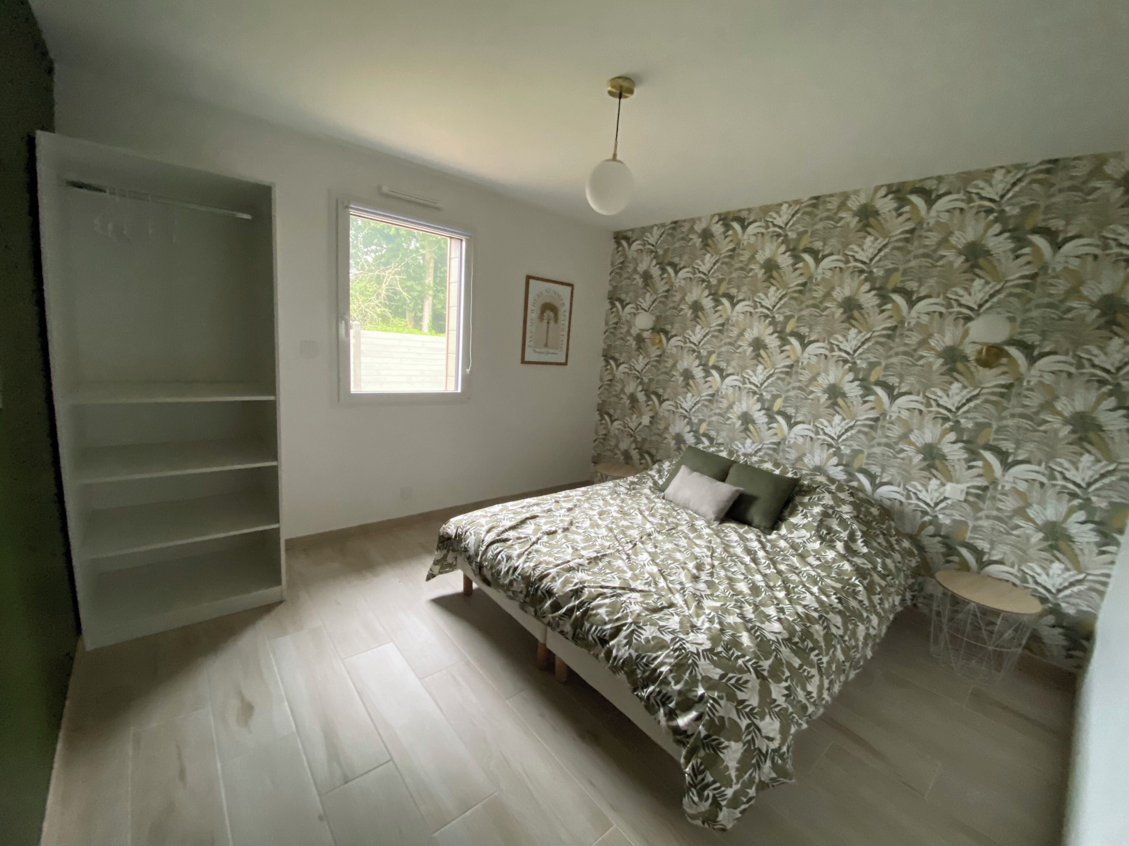 Ein Schlafzimmer mit Bett, Kleiderschrank, Fenster und blumigem Wandpapier.