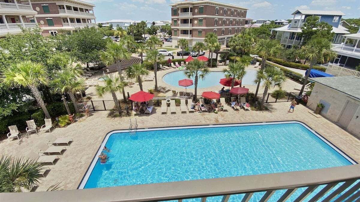Zona de piscina con sillones de descanso y sombrillas, rodeada de palmeras y un edificio con varios balcones.