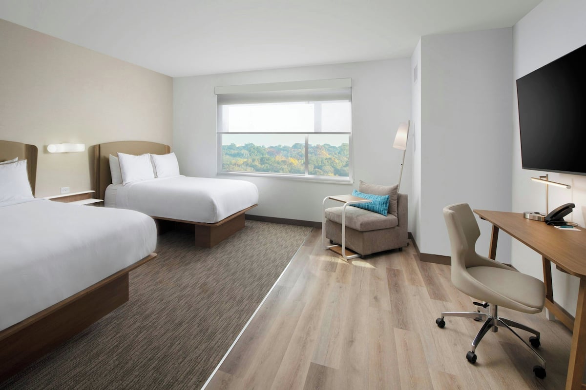 A hotel room with two beds, a desk, a chair, a TV, and a window with a view of trees.