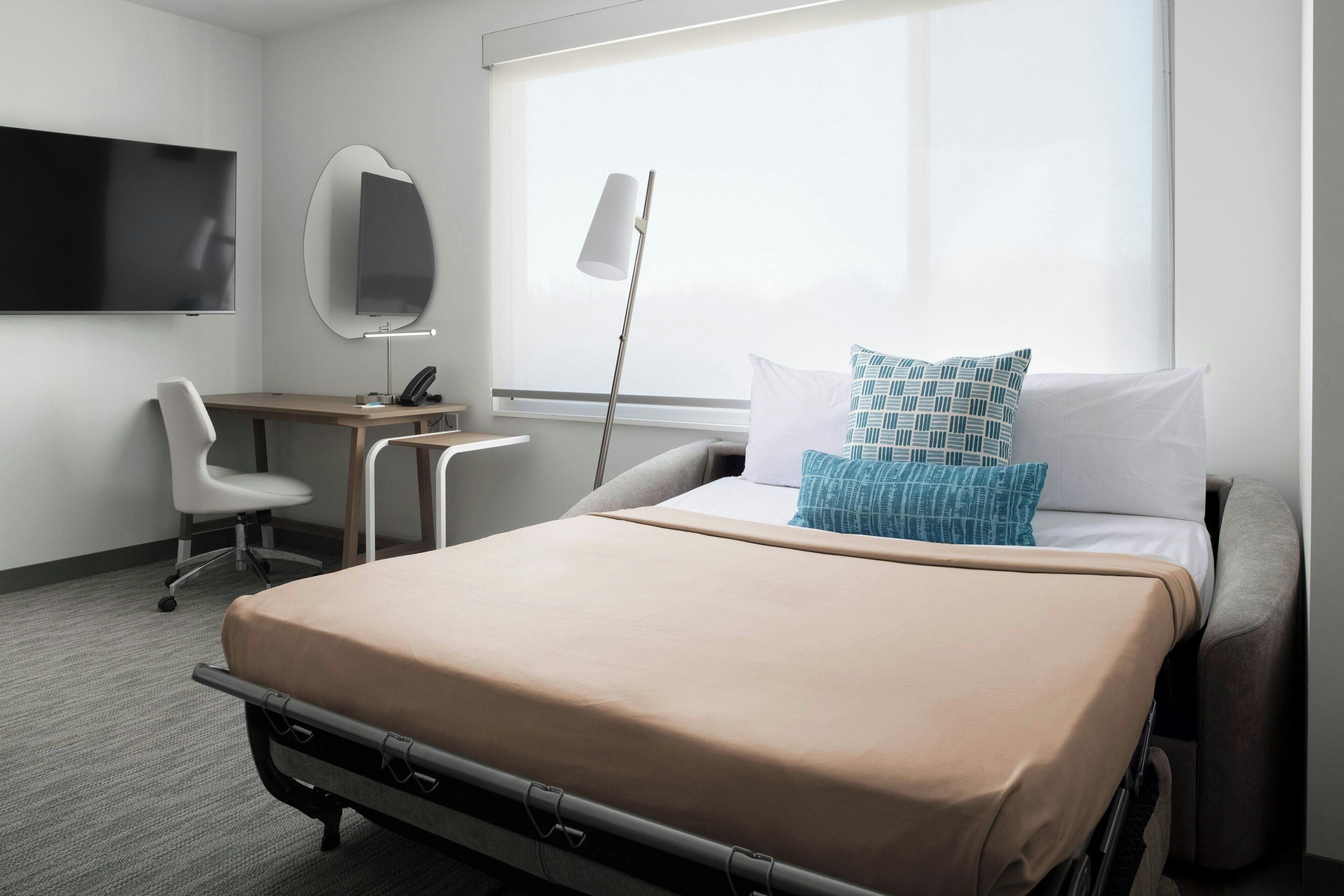 A hotel room with a bed, a desk, a chair, a TV, and a window.